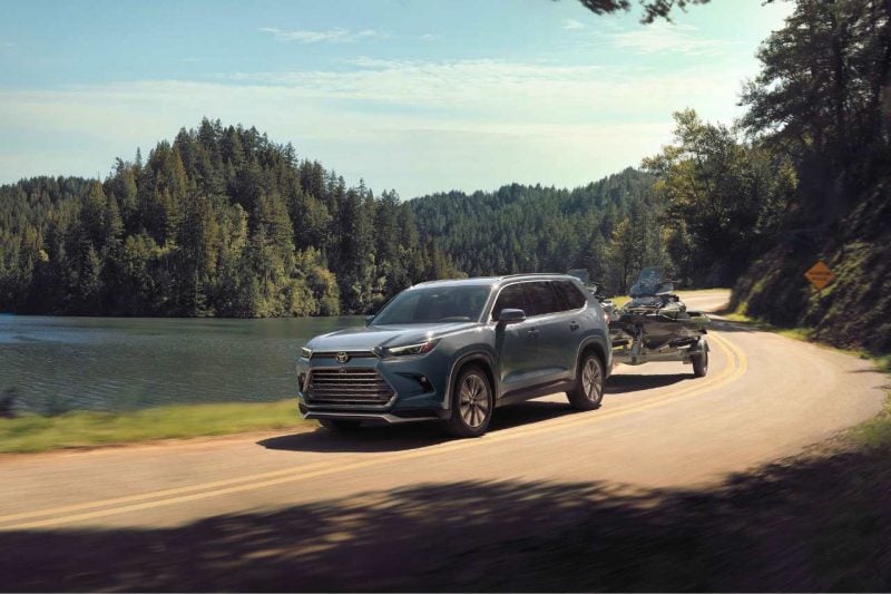 A 2024 Grand Highlander vehicle is towing a sports vehicle along a road.