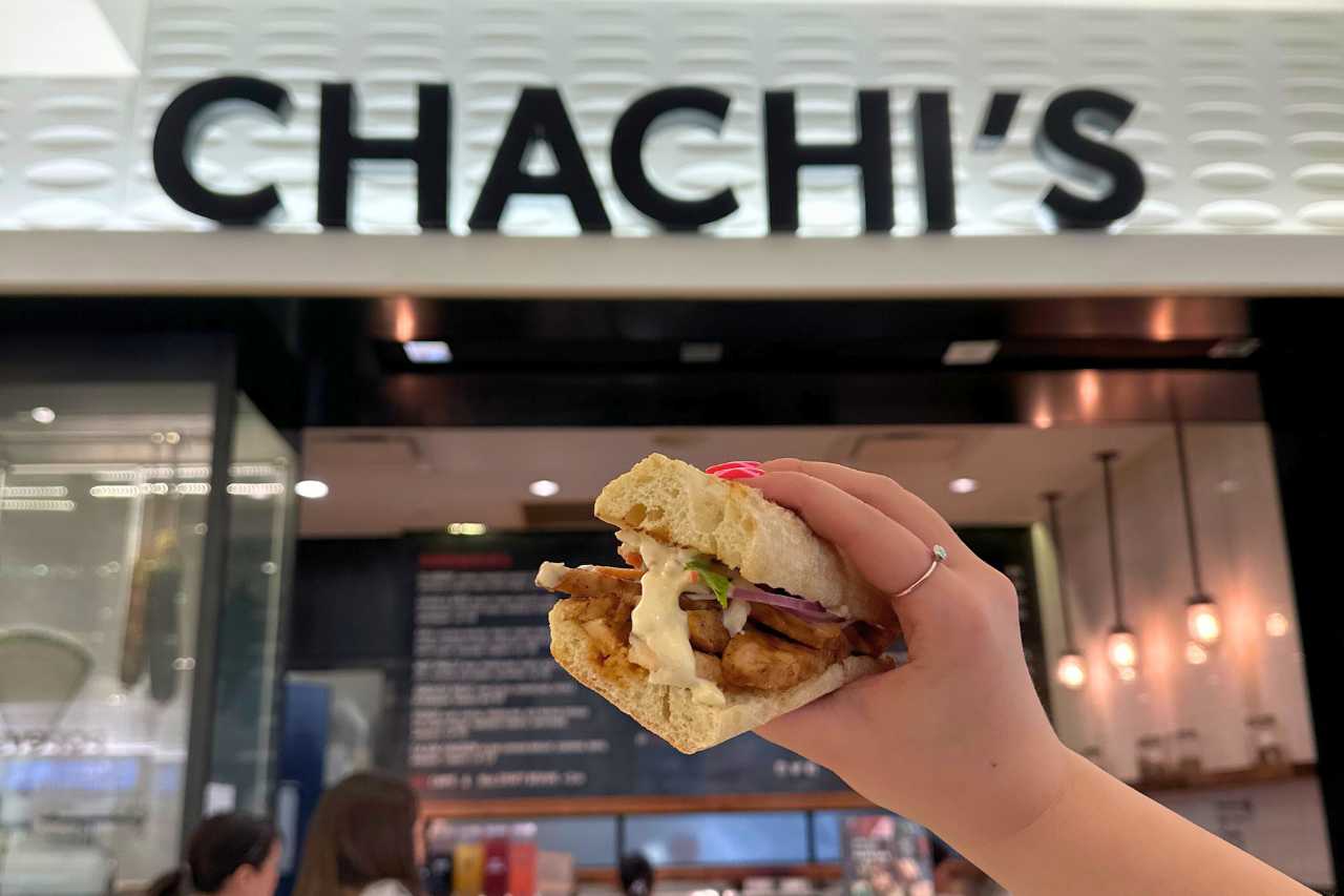 A woman holds up a Chicken Banh Mi sandwich in front of a Chachi's sandwich shop.