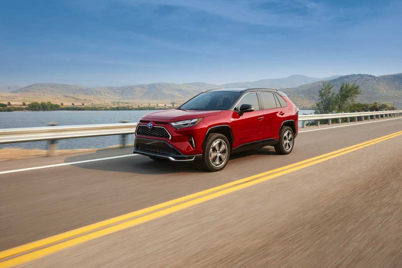 A red 2024 Toyota RAV4 Prime drives down a sunny highway next to a beach.