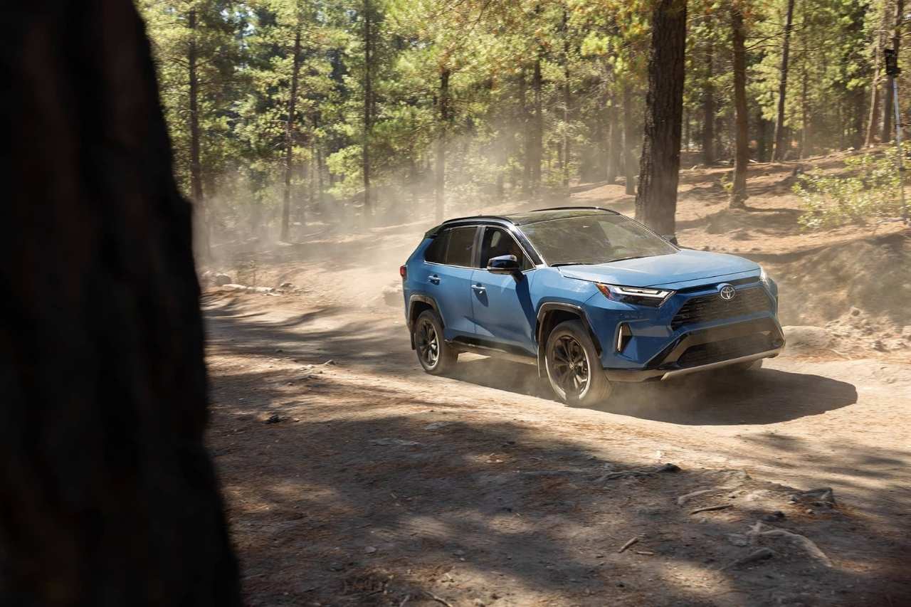 A 2024 Toyota RAV4 SUV in blue drives down a dirt road in a forest.