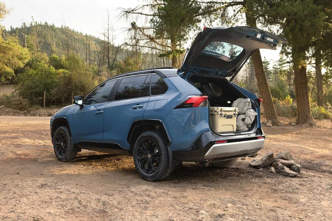A 2024 Toyota RAV4 in blue is parked in a forest with the trunk open, suitcases and camping gear packed into the back.
