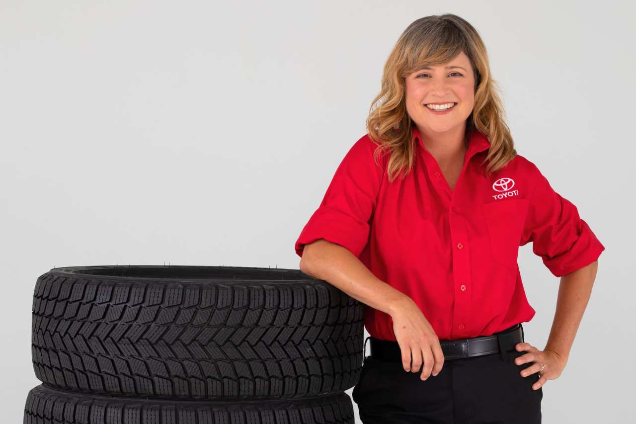 A female Toyota Tire Consultant smiles while standing against a pile of new tires.