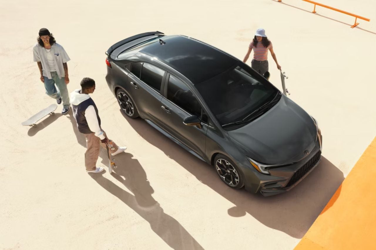 A few teenagers holding sports gear, like skateboards, walk up to their grey Corolla sedan.