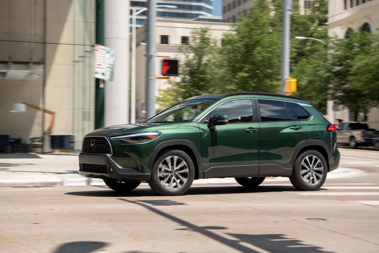 A green 2025 Toyota Corolla Cross SUV drives through a busy downtown city street.
