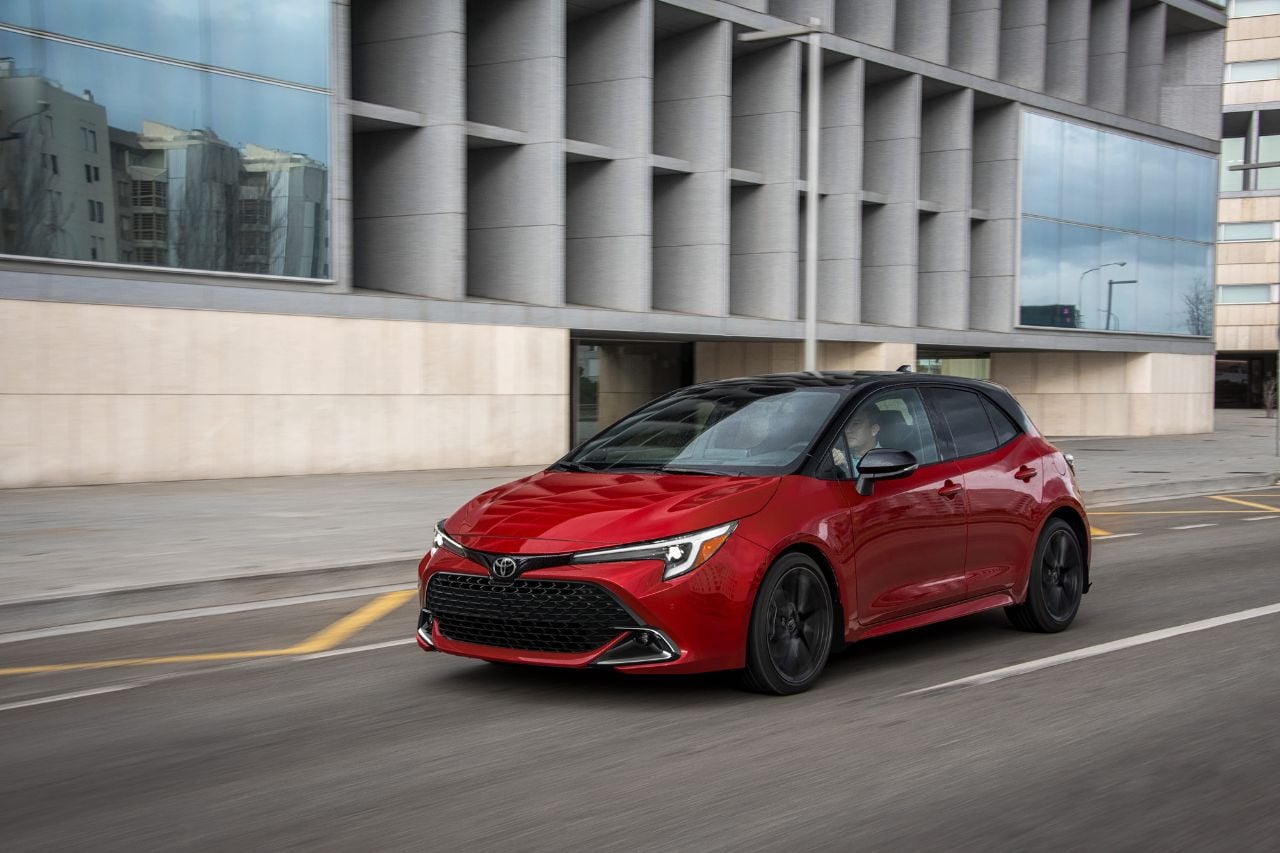A red hatchback car drives down city streets on a sunny afternoon.
