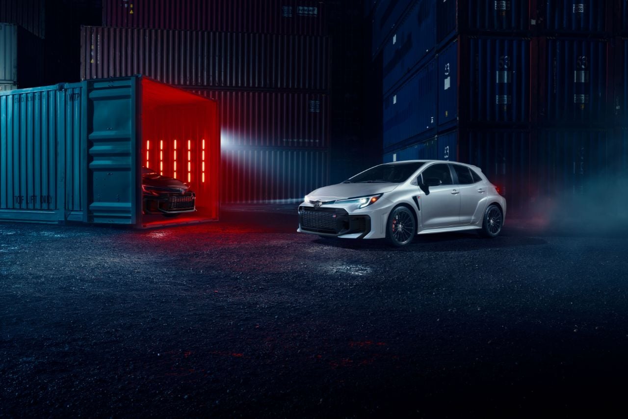 A white sports hatchback stands in a dark outdoor area, with a black vehicle sits in a shipping container illuminated in red lights next to it.