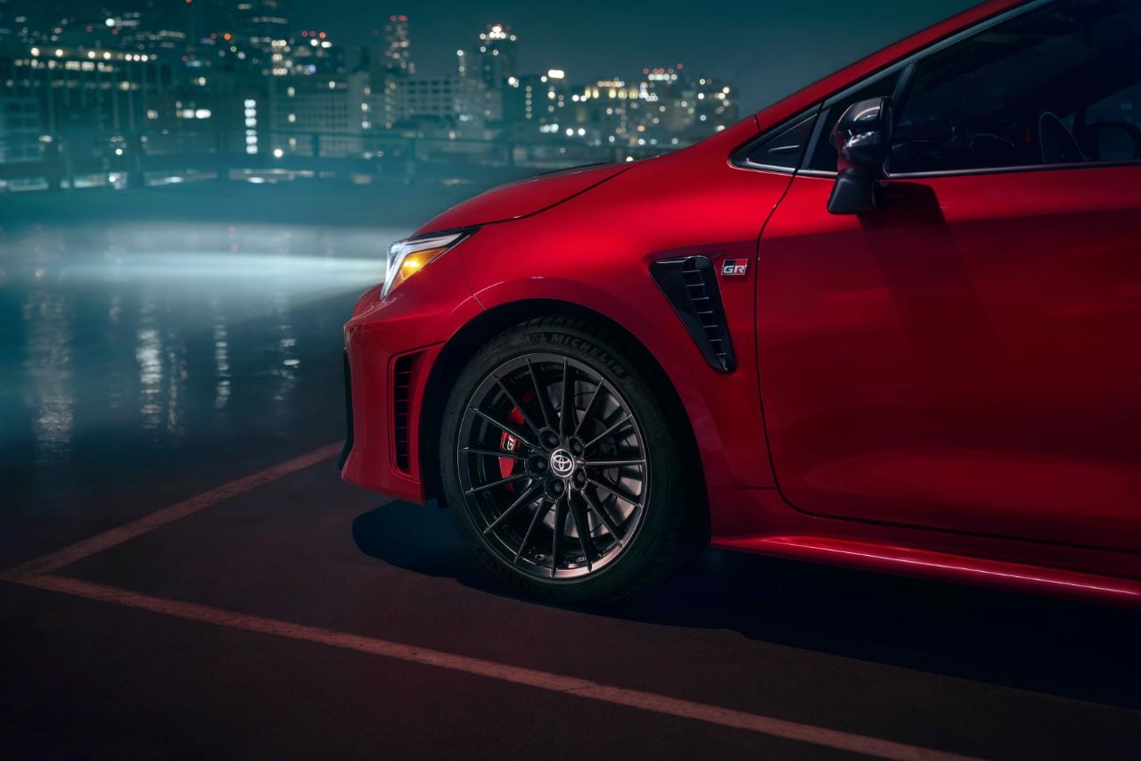 A red 2025 Toyota GR Corolla hatchback is parked with its headlights on, with a view of a city downtown in the background.