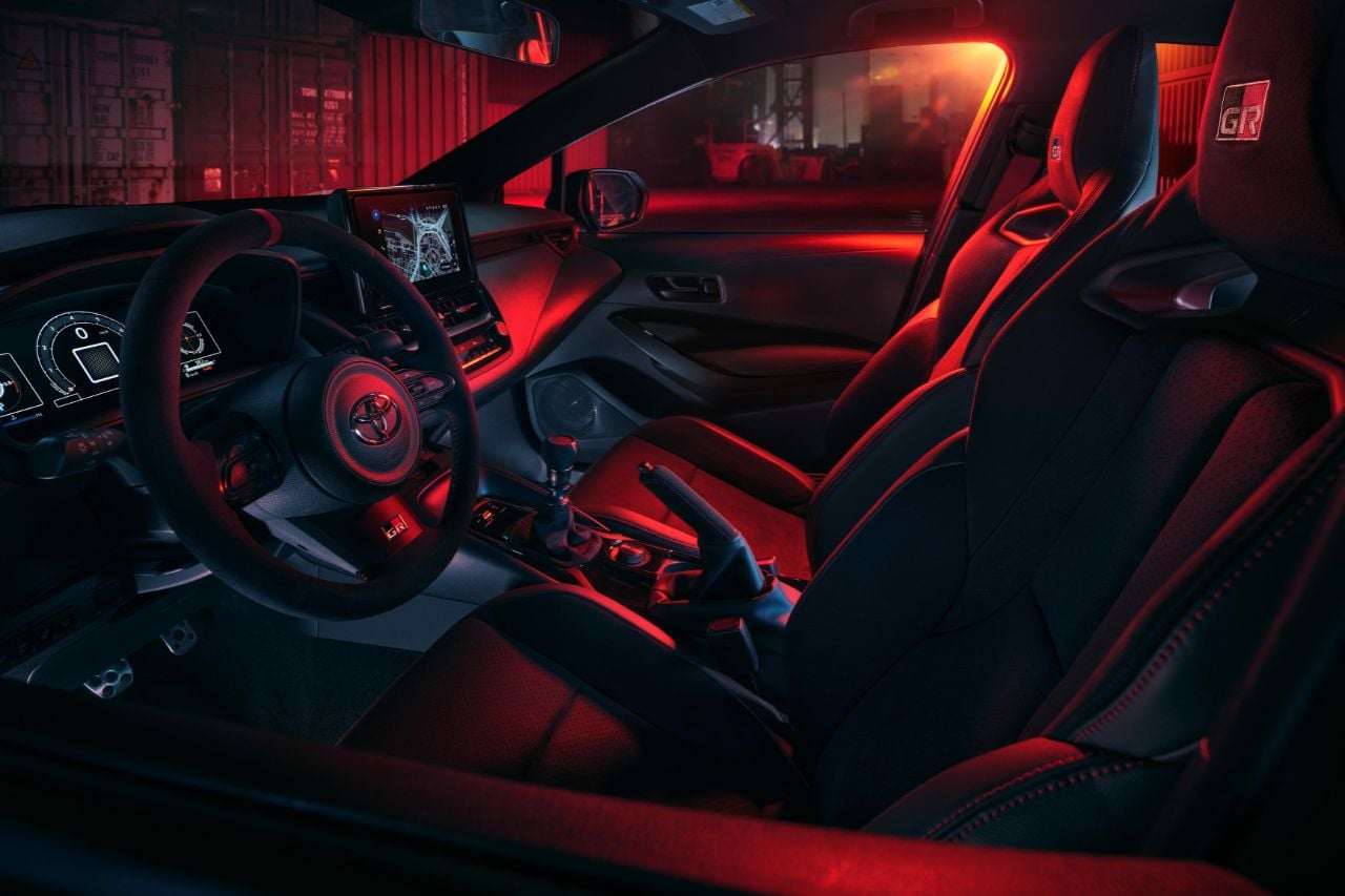 A red light illuminates the interior of a new GR Corolla sports hatchback, showing the steering wheel and dashboard.