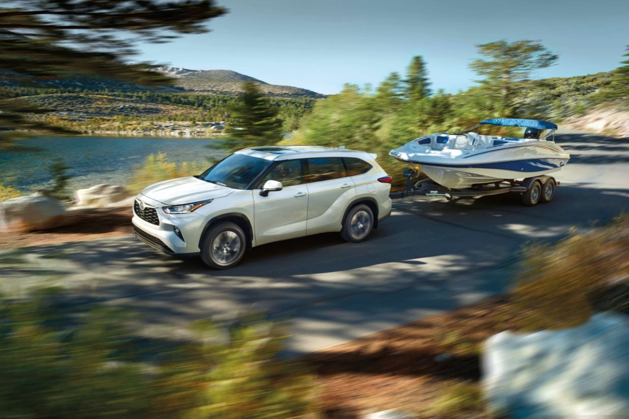 A white Highlander SUV drives down a backroad in the forest towing a speedboat on a sunny afternoon.