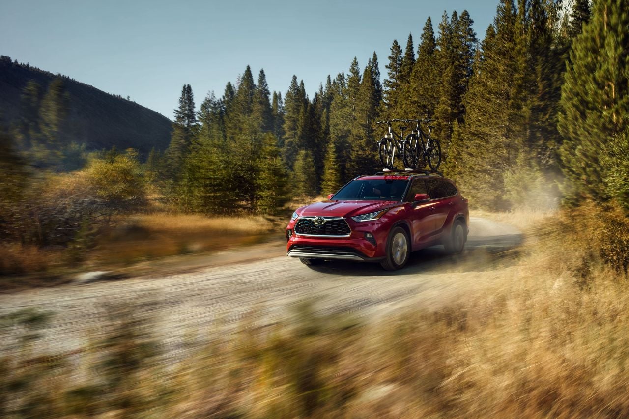 A 2025 Toyota Highlander SUV in a red colour barrels down a dirt road in the mountains with a roof rack carrying mountain bikes.
