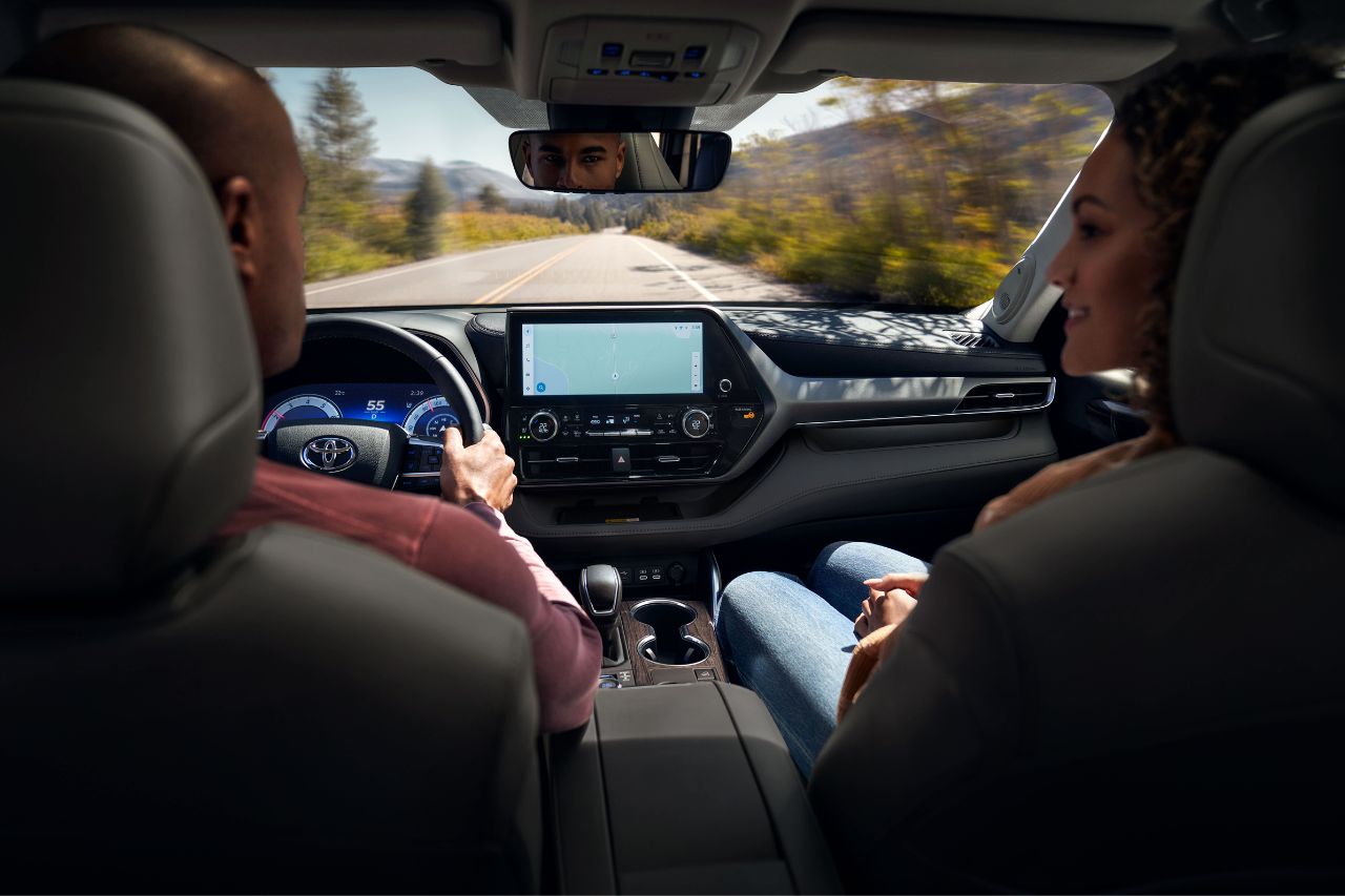 A man and woman drive a new Highlander SUV down a remote road, enjoying the leather interior and new tech features from Toyota.