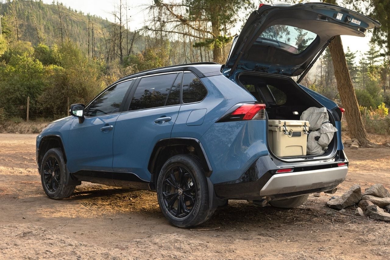 A blue RAV4 SUV is parked outside a forest camping ground with the trunk open, showcasing its cargo space.