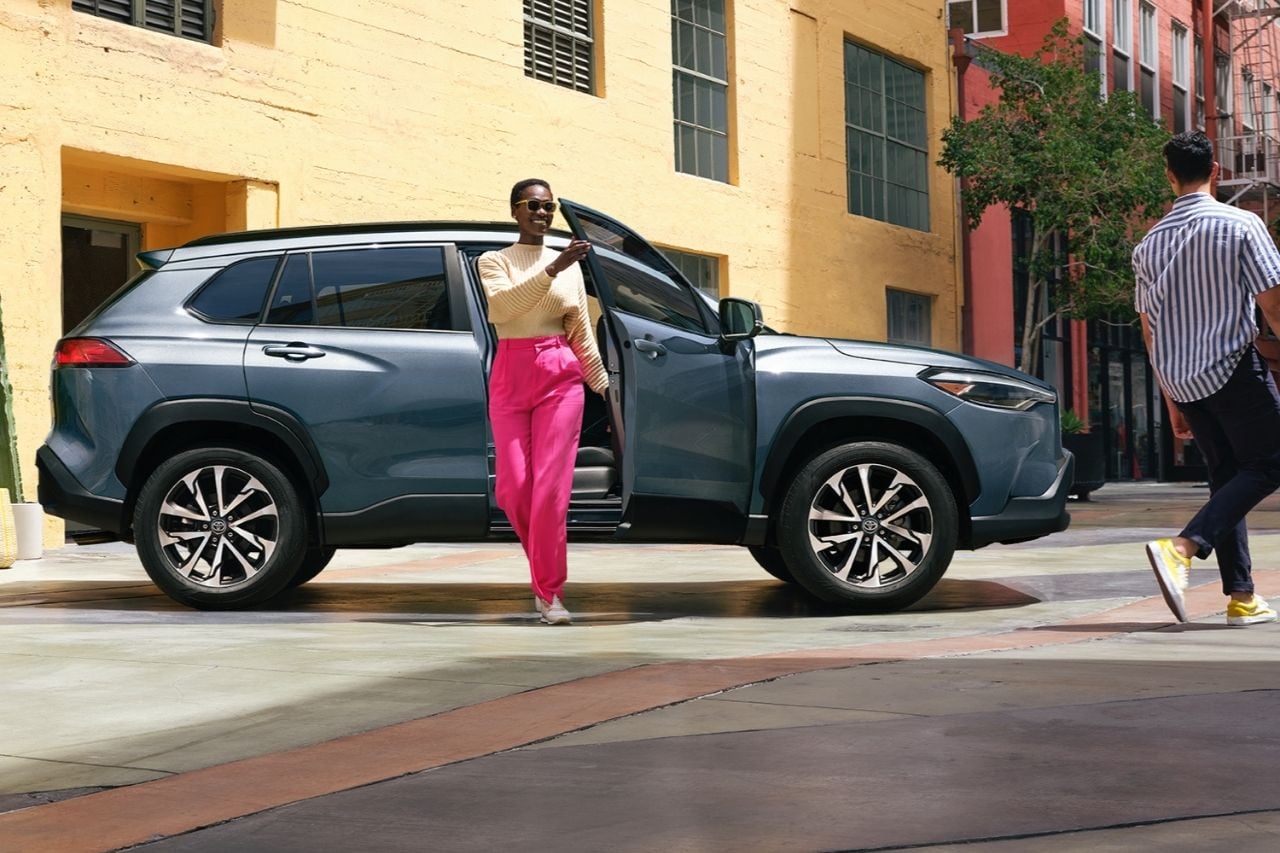 A shiny silver SUV is parked on a sunny stret, as an African American woman steps out of the vehicle wearing bright colours.