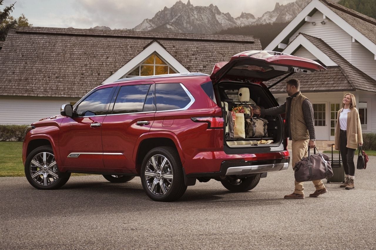 A large red SUV is parked outside a family home while a white couple load the trunk with suitcases.