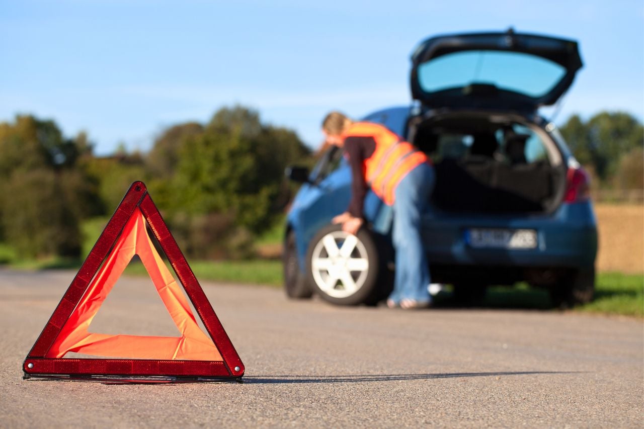 A young Caucasian woman has pulled off to the side of the road and is changing a tire. A reflective orange triangle is placed on the road for safety.