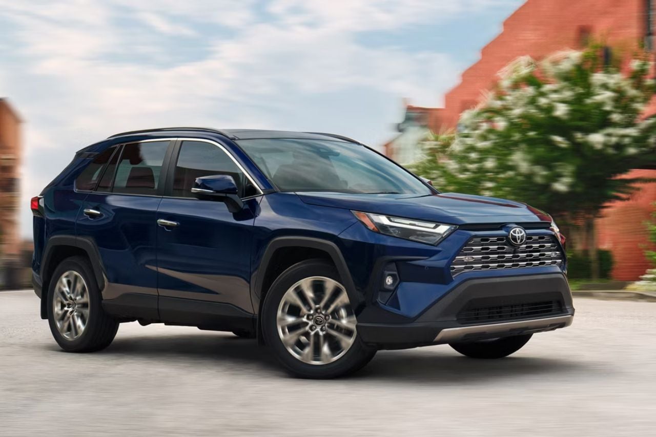 A brand-new 2025 Toyota RAV4 SUV in a deep blue colour drives down a city road on a bright, sunny afternoon.
