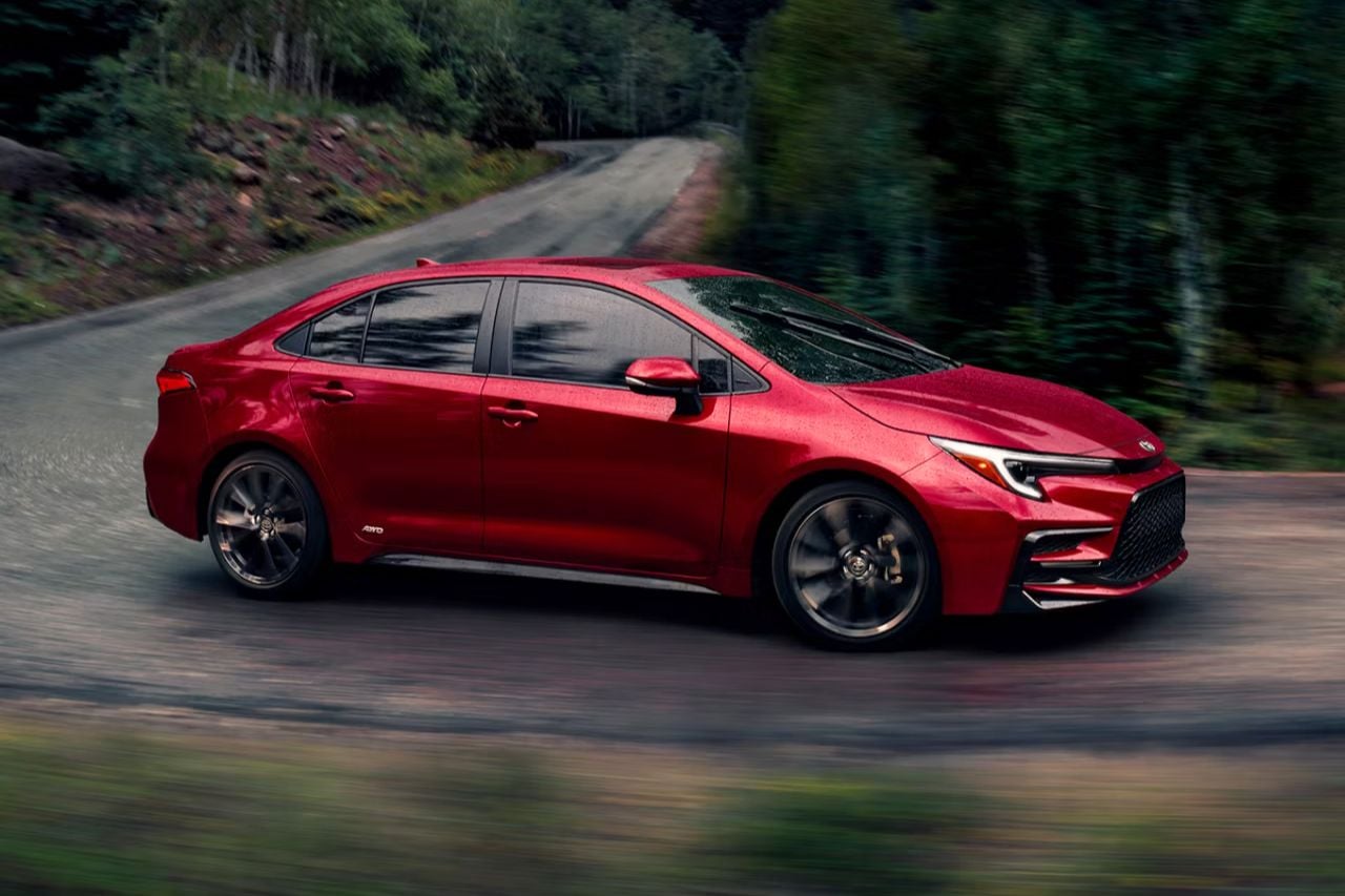 A brand new red Corolla sedan drives on a rainy, winding highway with trees in the background.
