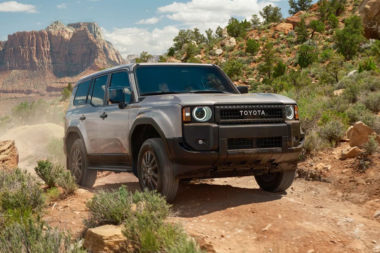 A Toyota Land Cruiser drives down a dusty, desert road on a sunny day.