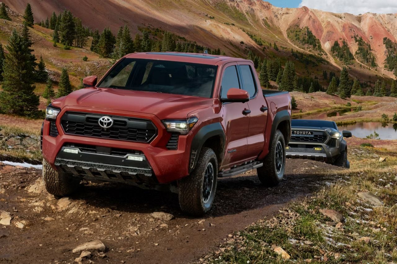 A red Toyota Tacoma truck drives down a dirt road with mountains in the background.