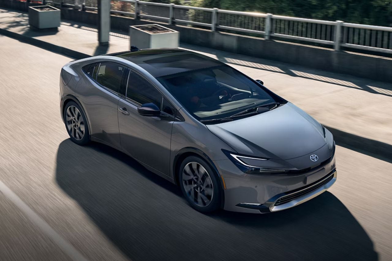 A grey 2026 Toyota Prius Plug-In Hybrid drives down a sunny highway road in Canada.