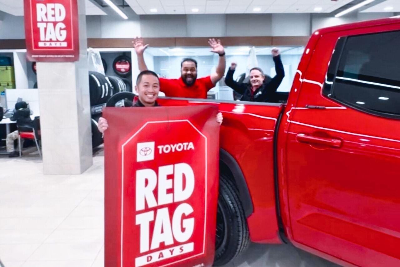 Sales team members at West Edmonton Mall Toyota smile and hold a "Red Tag Days" sign next to a red Toyota truck in the dealership showroom.