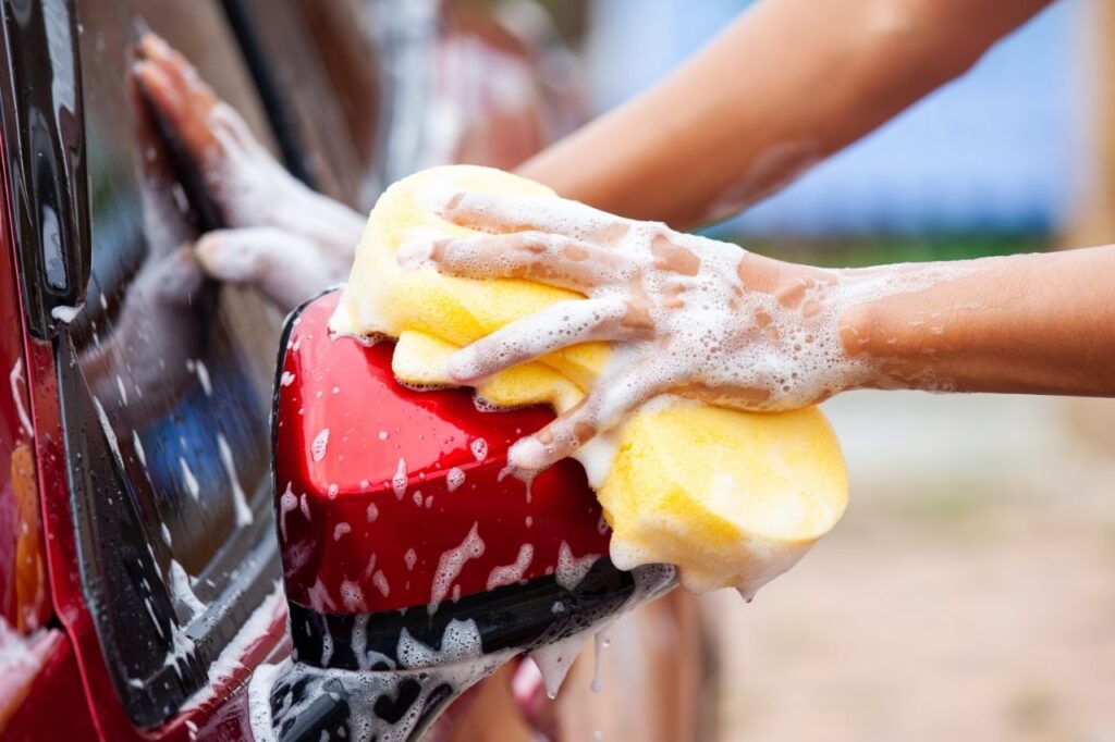 A person uses soap and a household sponge to DIY clean the exterior of their vehicle in their driveway.